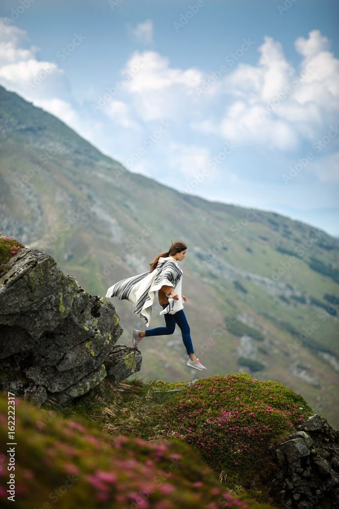Woman jumping on mountain peak rock. Young hipster woman hiker hiking on mountain trail. Hiker girl wrapping in warm poncho outdoor. Beautiful boho woman