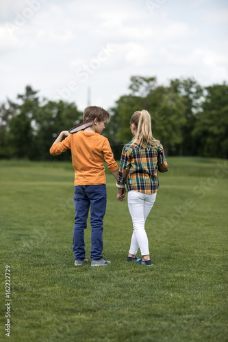 rear view boy and girl holding hands while standing on green field, boy holding badminton racquet on his shoulder