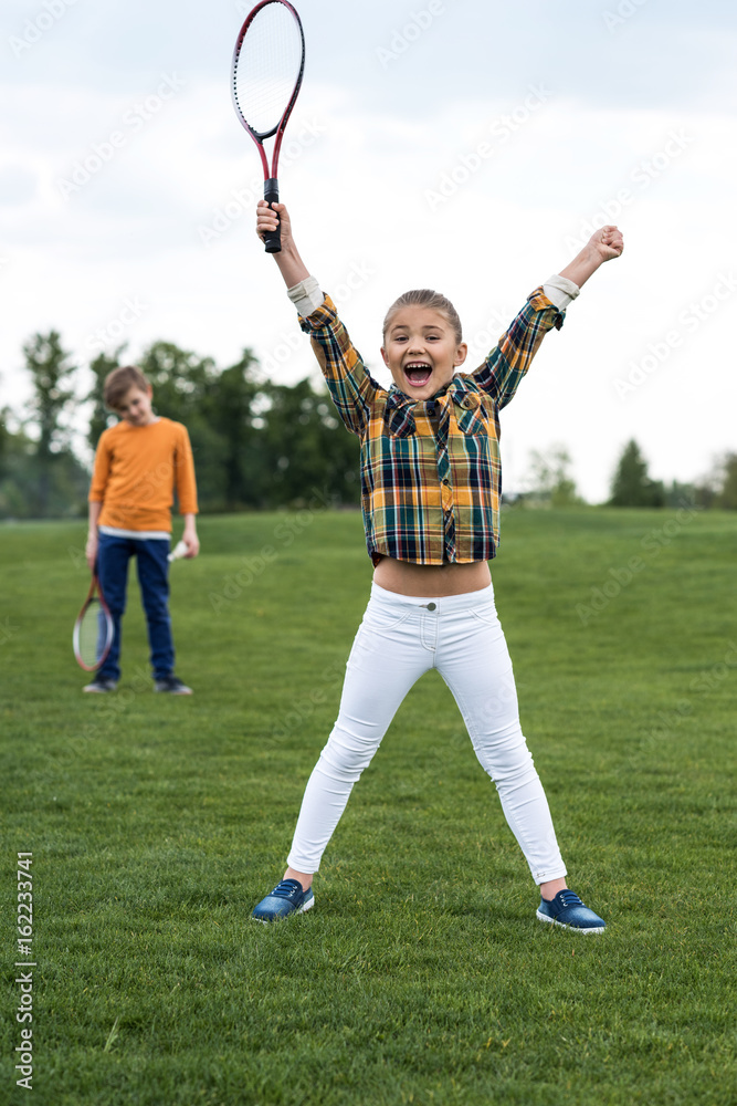 Obraz premium Happy child holding badminton racquet and triumphing while sibling standing behind