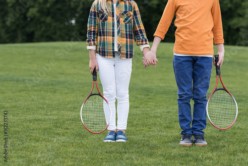 Cropped shot of siblings with badminton racquets holding hands and standing on green grass