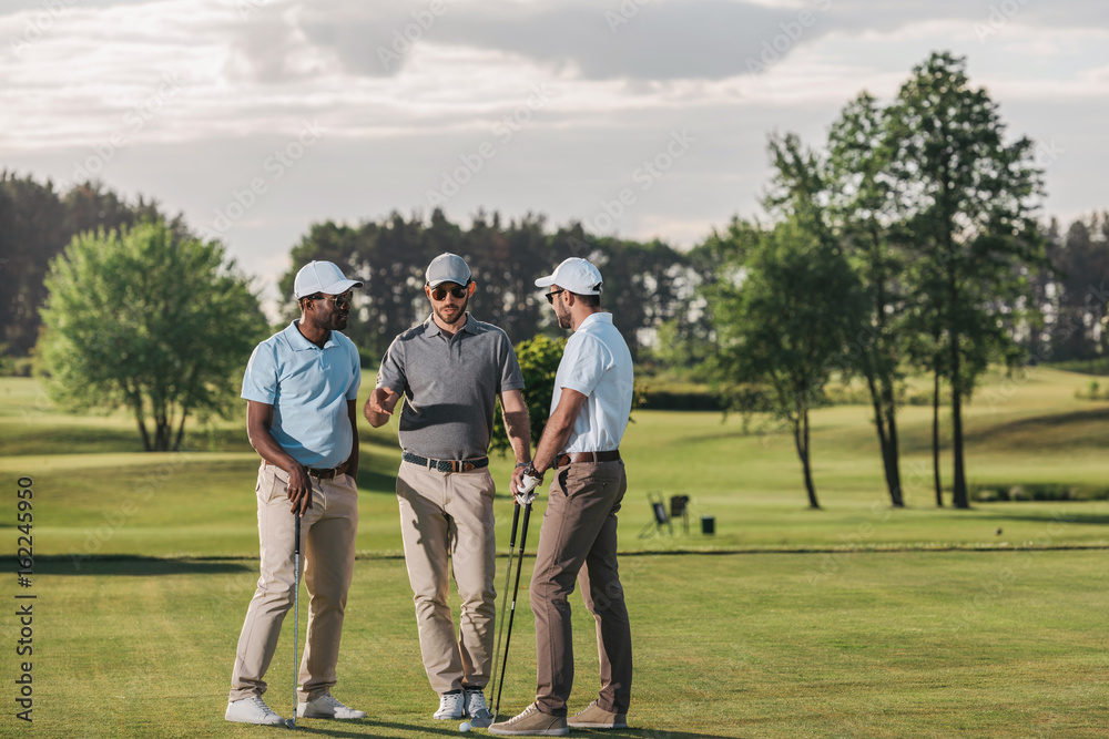 Multiethnic group of golfers holding clubs and talking while standing on green grass Multiethnic group of golfers holding clubs and talking while standing on green grass