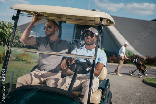 portrait of smiling man riding golf cart with friend sitting near by
