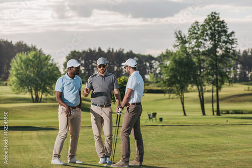 Multiethnic group of golfers holding clubs and talking while standing on green grass