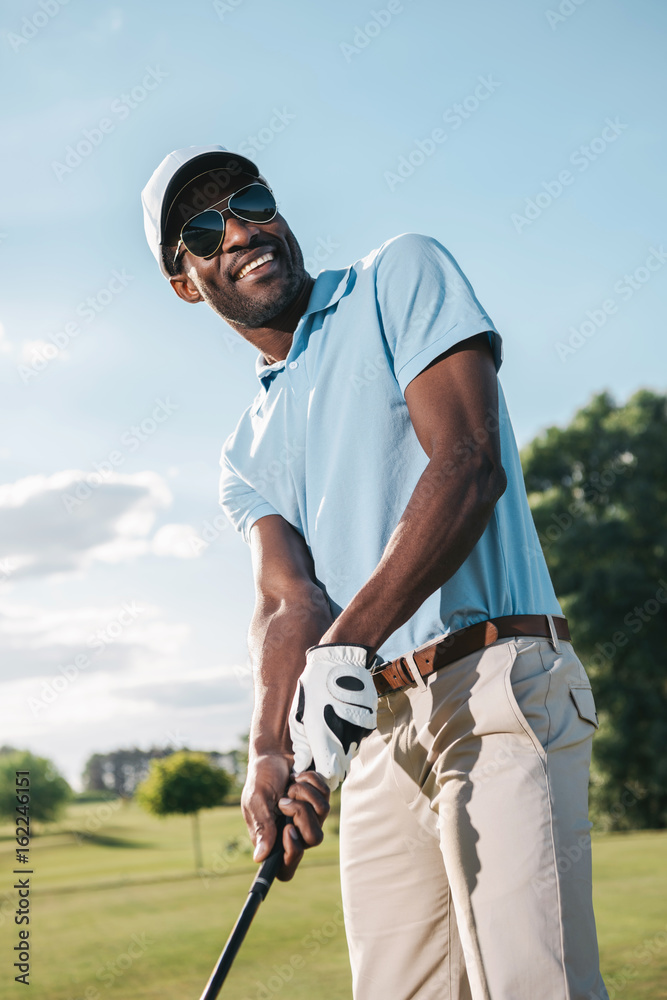 Smiling african american man in cap and sunglasses holding club and playing golf Smiling african american man in cap and sunglasses holding club and playing golf