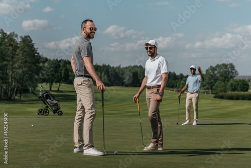 smiling friends talking while playing golf together on green at daytime