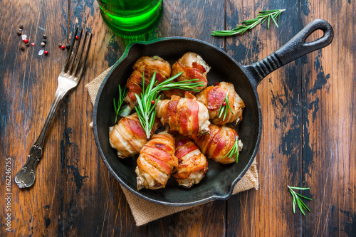 Oven roasted bacon wrapped chicken drumsticks in a black baking pan on the wooden rustic table, top view.