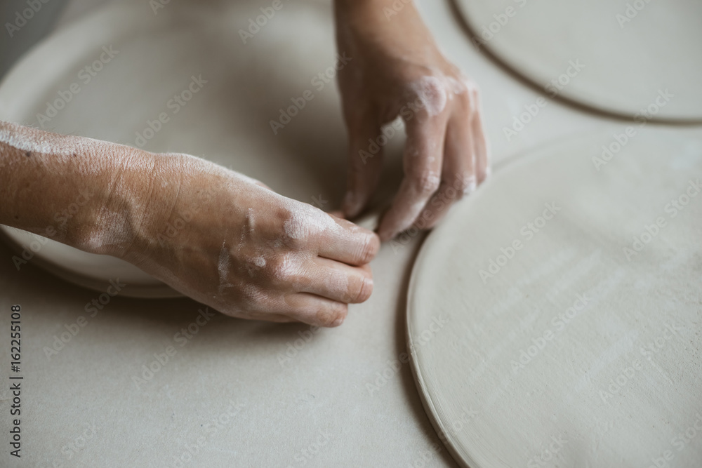 Woman's hands makes clay plates in pottery workshop, close up view; focus on stained hand Woman's hands makes clay plates in pottery workshop, close up view; focus on stained hand