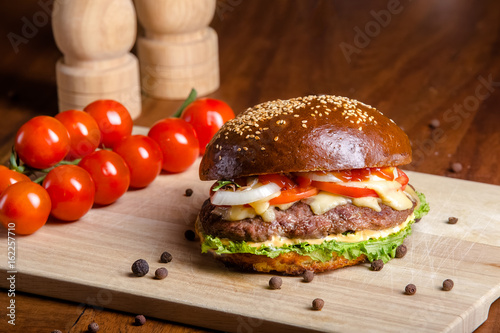 Toasted brown hamburger with slices of chopped red tomatoes, mozzarella cheese, tomato sauce, slice of fried bacon, juicy steak from pork or beef, greens and tomato wooden chopping board, whole champi photo