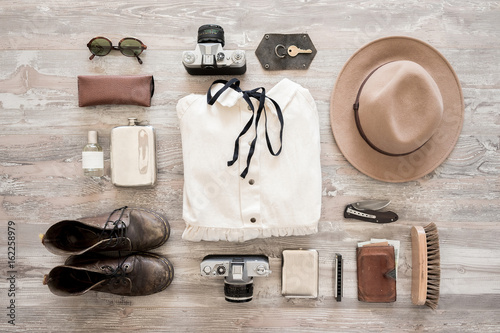 Preparation for traveling concept, dress, old shoes, jeans, beige hat, vintage cameras, leather bag, sunglasses, wallet, woman bra and another stuff on a white wooden background.
