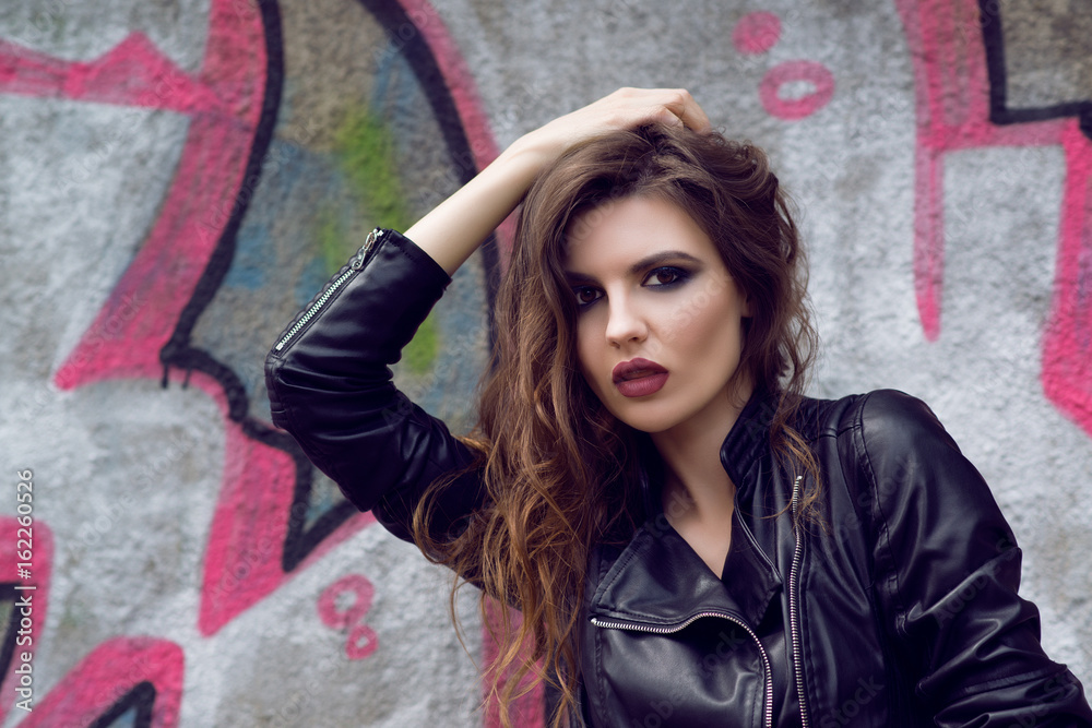 Portrait of the beautiful young woman with wavy brown hair posing outdoor, over graffiti background. Toned.