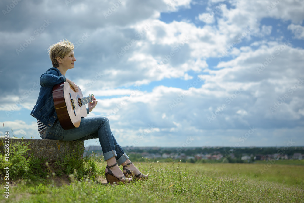 Obraz premium young woman with short blond hair sitting with guitar on the field in cloudy day
