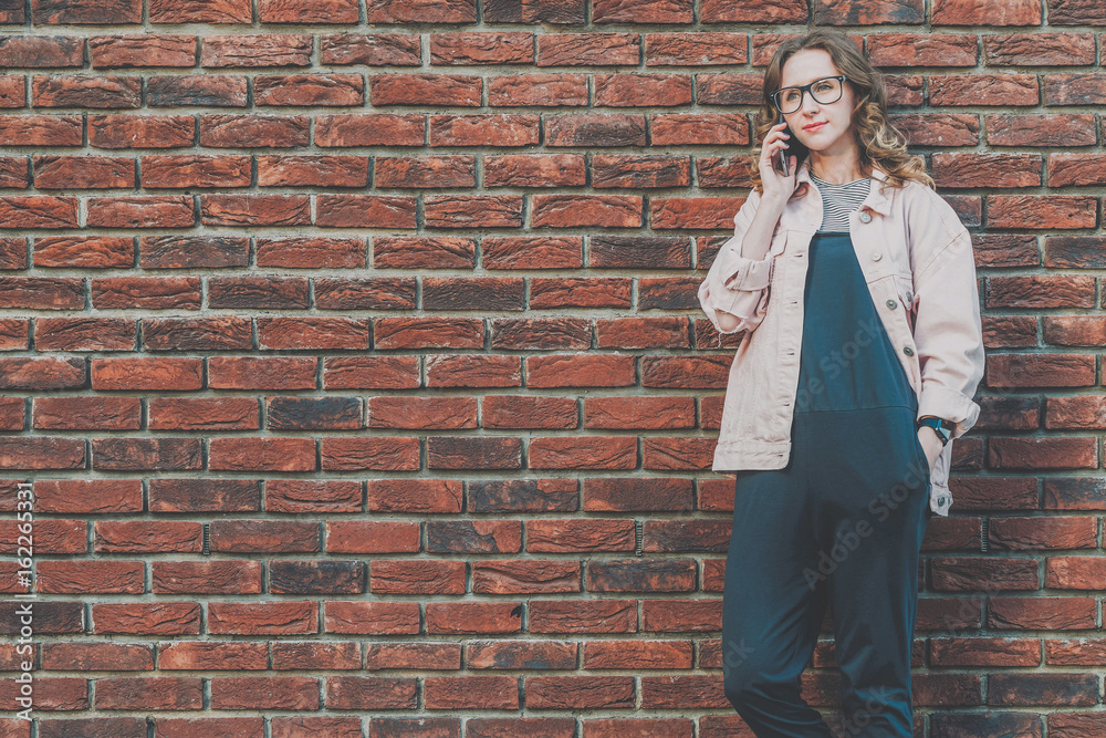 Young attractive hipster woman stands outside on a red brick wall background and talks on her cell phone. Young attractive hipster woman stands outside on a red brick wall background and talks on her cell phone.