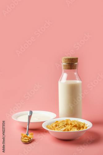 Corn flakes and yogurt in white bowl with milk bottle place on pink background