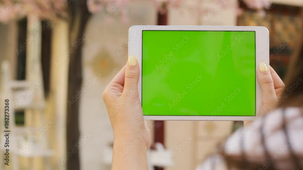 Woman using white tablet device computer with green screen touchscreen outdoors in the street. Sacura tree background. Close up. Chroma key. Shot over shoulder Woman using white tablet device computer with green screen touchscreen outdoors in the street. Sacura tree background. Close up. Chroma key. Shot over shoulder