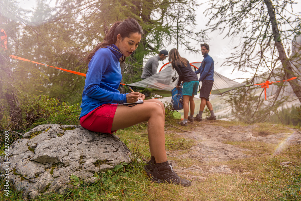 Obraz premium Brunette woman writing personal diary while friends setting hanging tent camping. Group of friends people summer adventure journey in mountain nature outdoors. Travel exploring Alps, Dolomites, Italy.