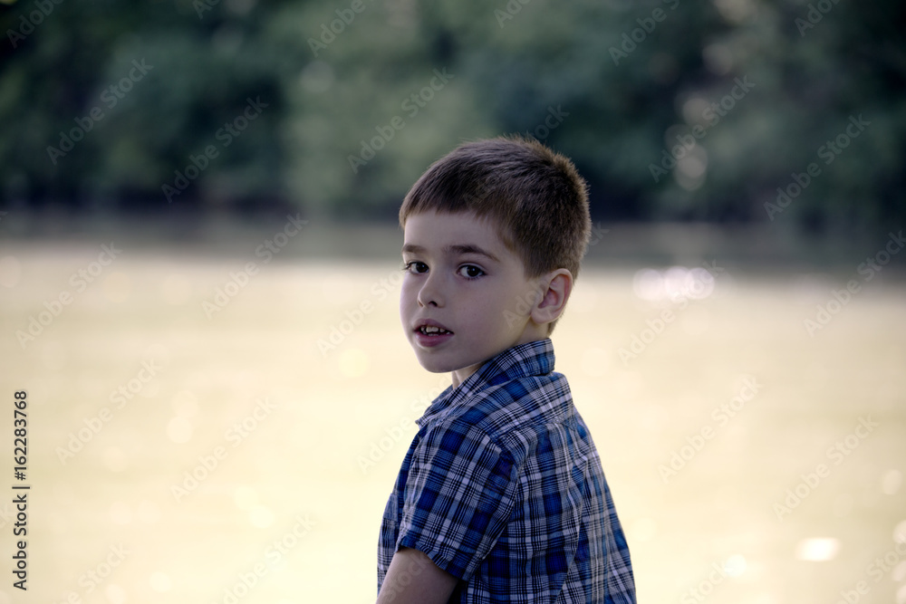 Smiling boy looks into camera on pier near pond. Little kid thinks and looks at the pond. Dream positive atmosphere, sunny day.