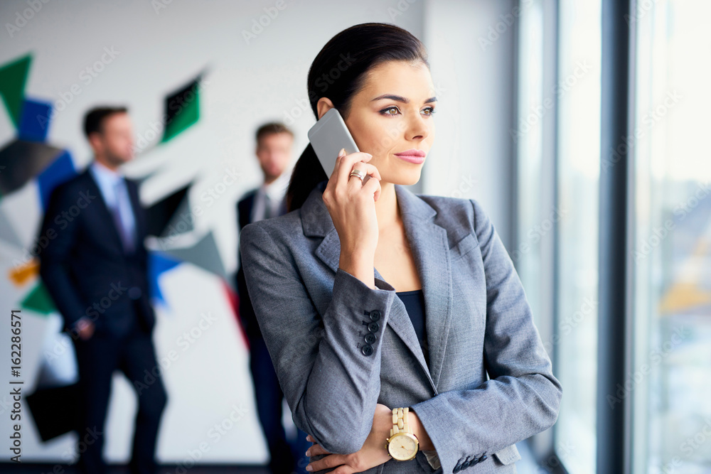 Portrait of young businesswoman talking on phone with two coworkers in the background Portrait of young businesswoman talking on phone with two coworkers in the background