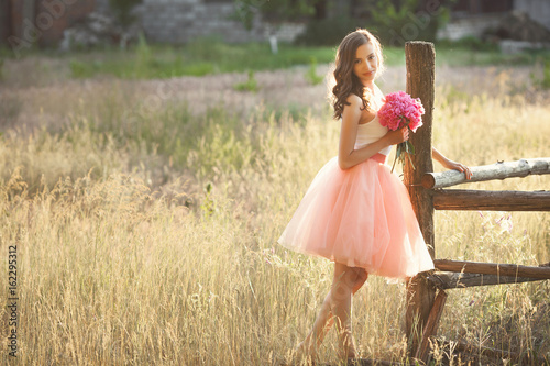 Beautiful young girl with pions on the nature otdoors in the sunshine. Pretty woman outside with flowers in the pink skirt photo