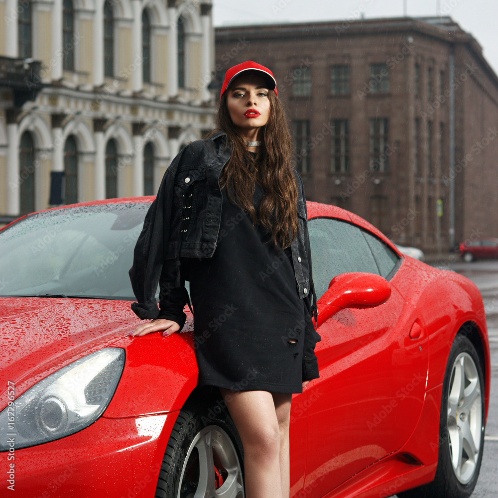 Fototapeta premium Glamorous sexy fashionable woman wearing sweetshot, jacket and red cap with long brown hair standing against red sport car at city street on overcast day