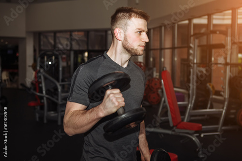 Handsome man doing biceps lifting with dumbbell on bench in a gym