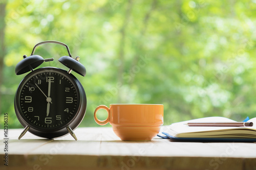 Man's hands holding a cup of coffee with notebook, pen and alarm clock on a Desktop from wooden plank in natural background.