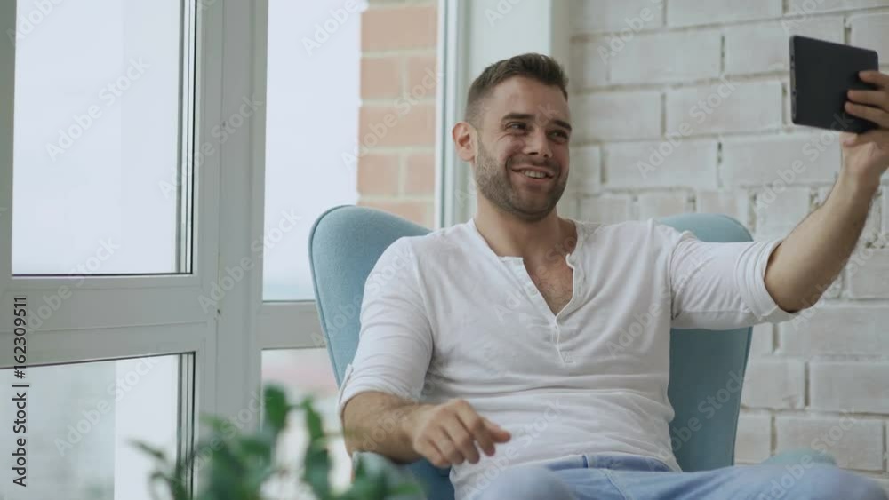 Young happy man have online video chat using digital tablet computer sitting on balcony in modern loft apartment