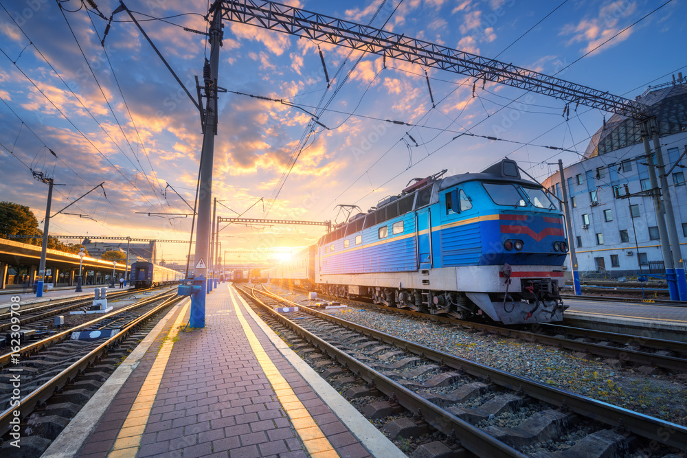 Obraz premium Blue train at the railway station at sunset. Amazing industrial landscape with old locomotive, old buildings, rails and colorful sunset sky with clouds. Train on railroad. Beautiful railway platform