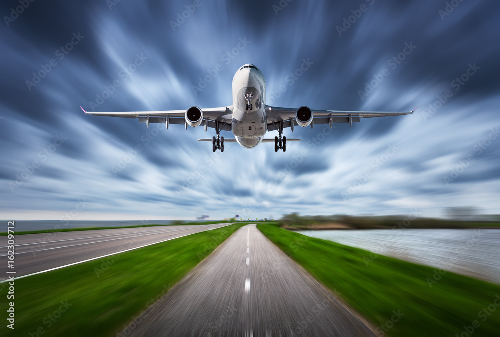 Obraz premium Airplane and road with motion blur effect. Landscape with white passenger airplane is flying in the cloudy sky over the asphalt road. Blurred. Passenger airplane is landing. Commercial plane. Aircraft