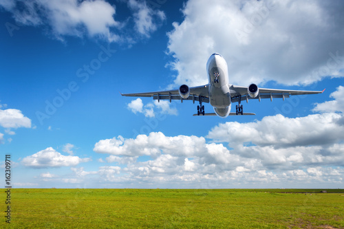 Beautiful airplane. Landscape with big white passenger airplane is flying in the blue sky with clouds over green grass field in summer. Passenger airplane is landing. Commercial plane. Aircraft