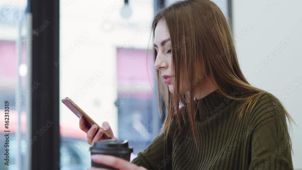 Pretty young woman in a stylish sweater sits in the café, drinks coffee, texts the messages and smiles happily. Cheerful mood, being happy. Having fun, daily routine. Pretty young woman in a stylish sweater sits in the café, drinks coffee, texts the messages and smiles happily. Cheerful mood, being happy. Having fun, daily routine.