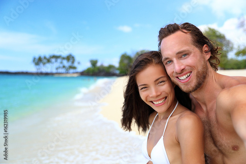 Happy beach selfie couple in love on Hawaii holidays. Healhty lifestyle young multiracial people smiling in swimwear on tropical vacation.