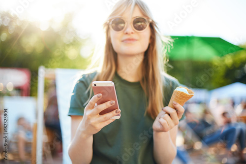 Woman eating icecream using smartphone. Portrait of happy girl with ice cream looking at camera browsing through social media or messaging her friends enjoying summer in the city park wearing shades