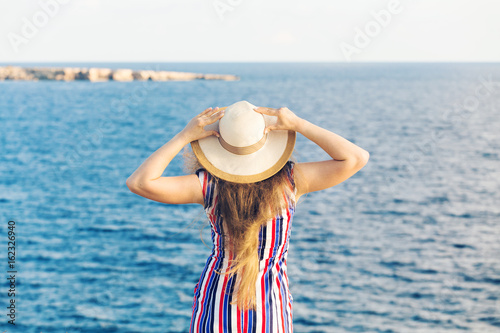 Young woman on the beach near the sea wearing dress and hat, rear view