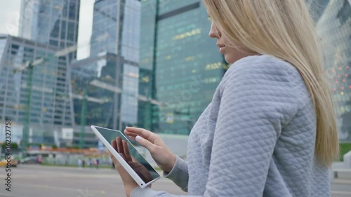 A young blond girl uses a tablet on a background of skyscrapers downtown. photo