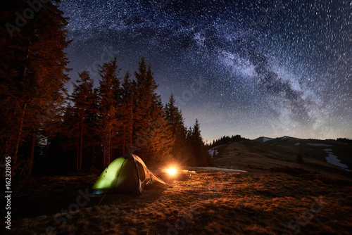 Night camping. Illuminated tent and campfire near forest under beautiful night sky full of stars and milky way