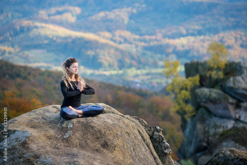 Sporty slim female is practicing yoga, meditating on the top of the mountain. Autumn forests, rocks and hills on the background