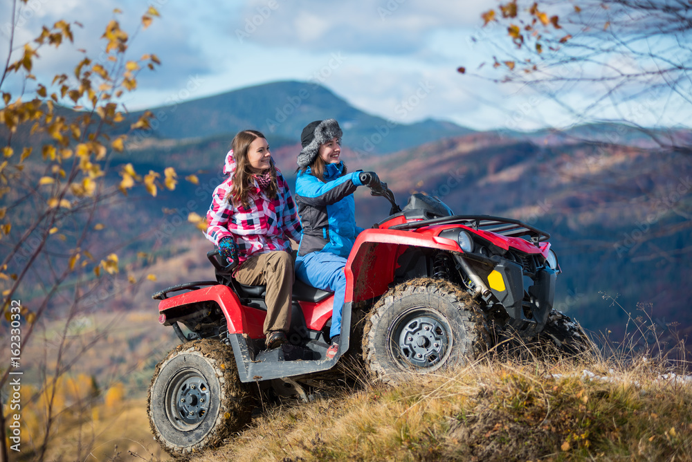 Fototapeta premium Happy females ATV driver in winter clothing on snowy hills on the background of mighty mountains and trees with yellow leaves