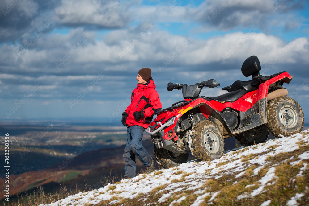 Atv quad bike near man in winter clothes looks into the distance on snowy mountain slope. On the background blue cloudy sky, mountains and the town in the valley with copy space