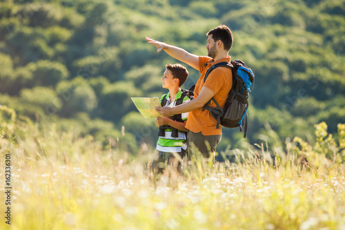 Father and son are hiking in nature in summer. They are looking at map.