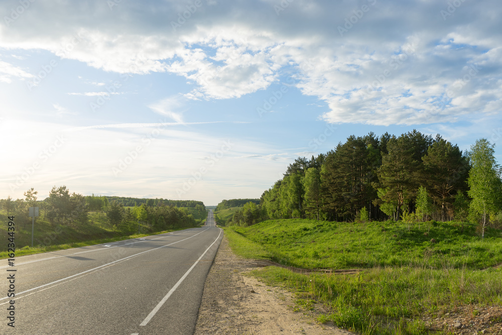 Vanishing straight highway with forest along in backlit at sunset
