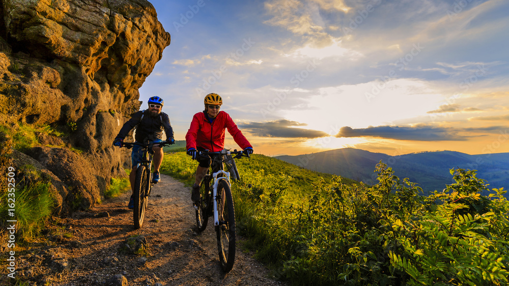 Naklejka premium Mountain biking women and man riding on bikes at sunset mountains forest landscape. Couple cycling MTB enduro flow trail track. Outdoor sport activity.