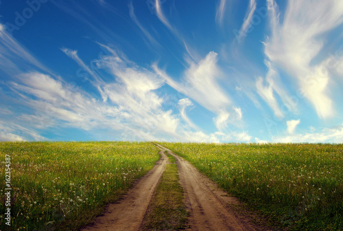 Rural road through green fields against the background of unusual sky