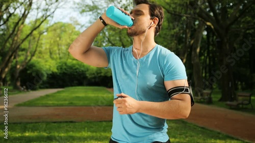 Attractive fit man drinking water from plastic botle after jogging, running, fitness training in park. Refreshing after workout. Looking at the camare. Portrait. Close up photo