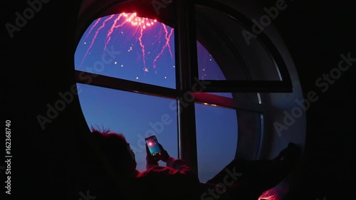 Girl taking fireworks video with a smartphone. Silhouette in a round loft window