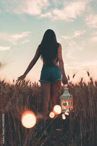 Giovane ragazza cammina tra le spighe di grano mentre tiene in mano la lanterna luminosa, al tramonto photo