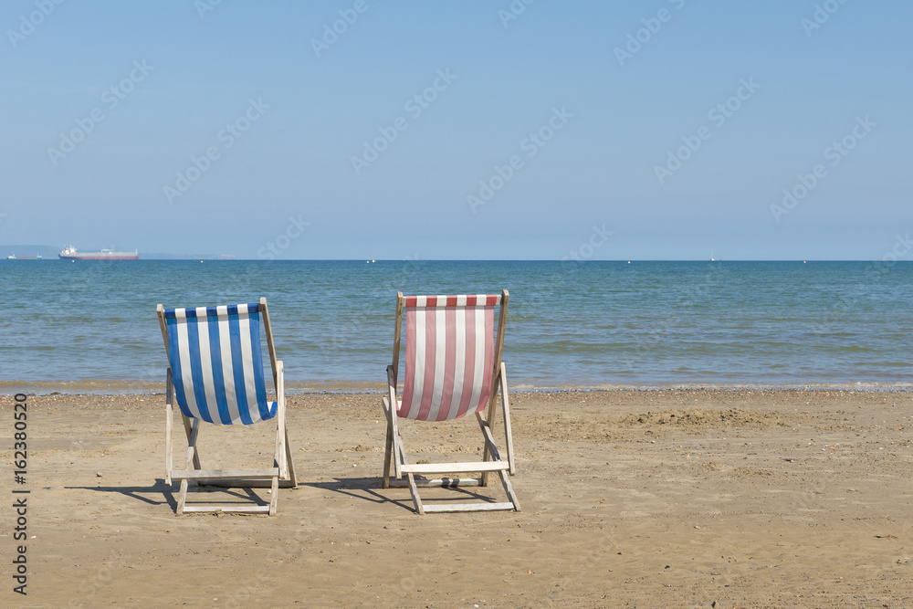 Two empty linen beach chairs one blue and one red in a sea landscape on the beach of Weymouth of the United Kingdom, facing the sea on a sunny day Two empty linen beach chairs one blue and one red in a sea landscape on the beach of Weymouth of the United Kingdom, facing the sea on a sunny day
