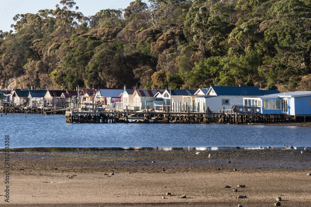 Hobart, Australia - March 19. 2017: Tasmania. Closeup of row of small beach houses built over blue river water at Cornelian Bay. Green tree vegetation in back. In front, beach. Hobart, Australia - March 19. 2017: Tasmania. Closeup of row of small beach houses built over blue river water at Cornelian Bay. Green tree vegetation in back. In front, beach.