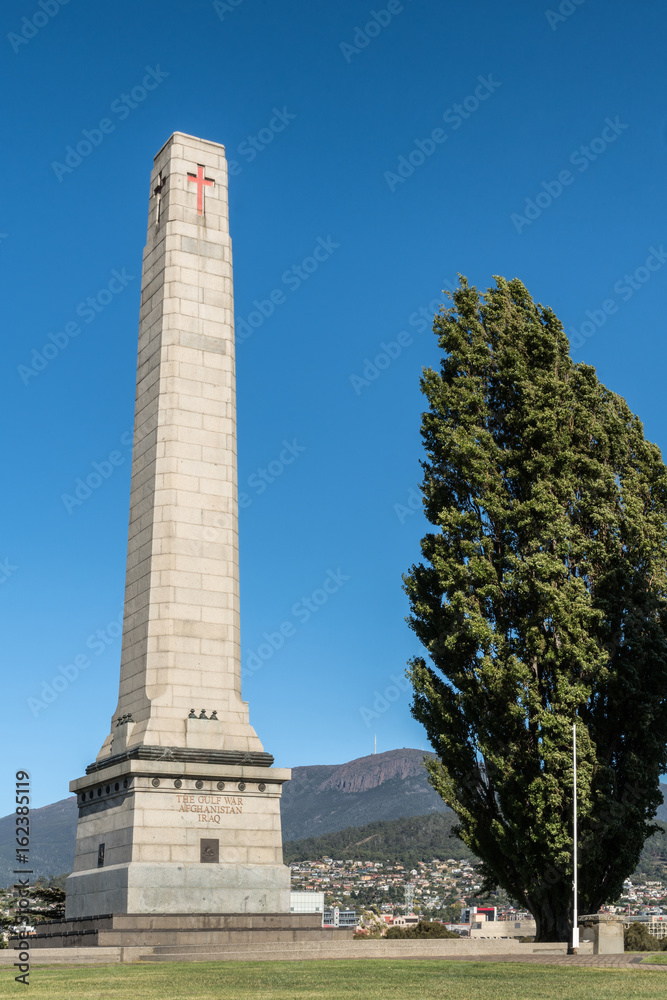 Hobart, Australia - March 19. 2017: Tasmania. Closeup of tall white stone Cenotaph war memorial and tree on green hill under blue sky. Gulf War, Iraq, Afghanistan side. City in back on mountain slope.