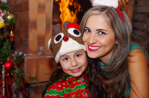 Happy mother and daugher at christmas, litle girl wearing a deer hat and mom a christmas hat, with an indoor chimmey background