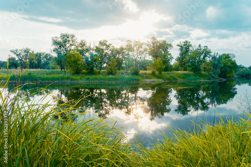 Calm beautiful scenic landscape with blue river, green trees and reflecting in water with cloudy sky. Magical sunset over the river in rural terrain. Natural, wild landscape.
